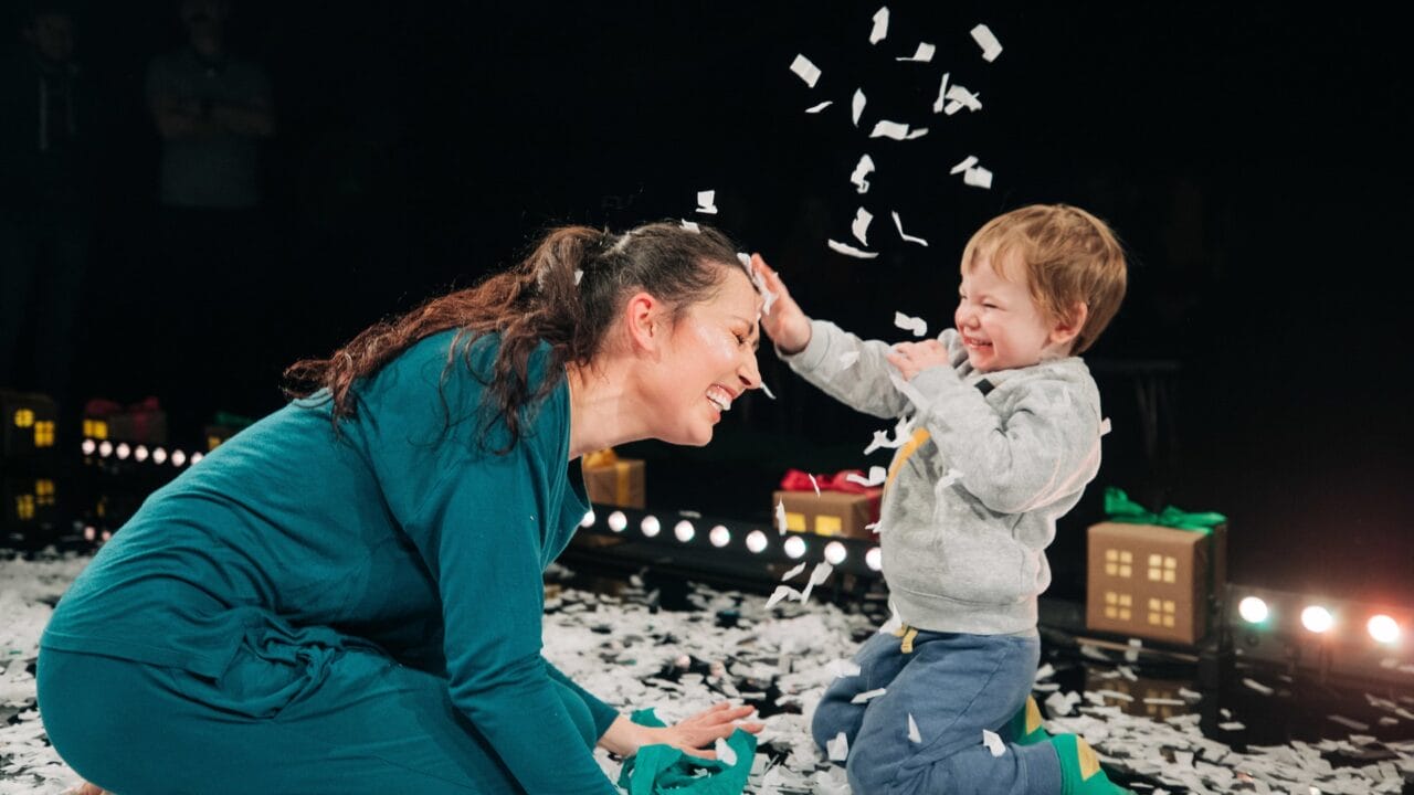 A woman kneels and smiles as a young child playfully throws confetti in the air; small paper pieces cover the floor and nearby props.