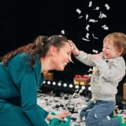 A woman kneels and smiles as a young child playfully throws confetti in the air; small paper pieces cover the floor and nearby props.