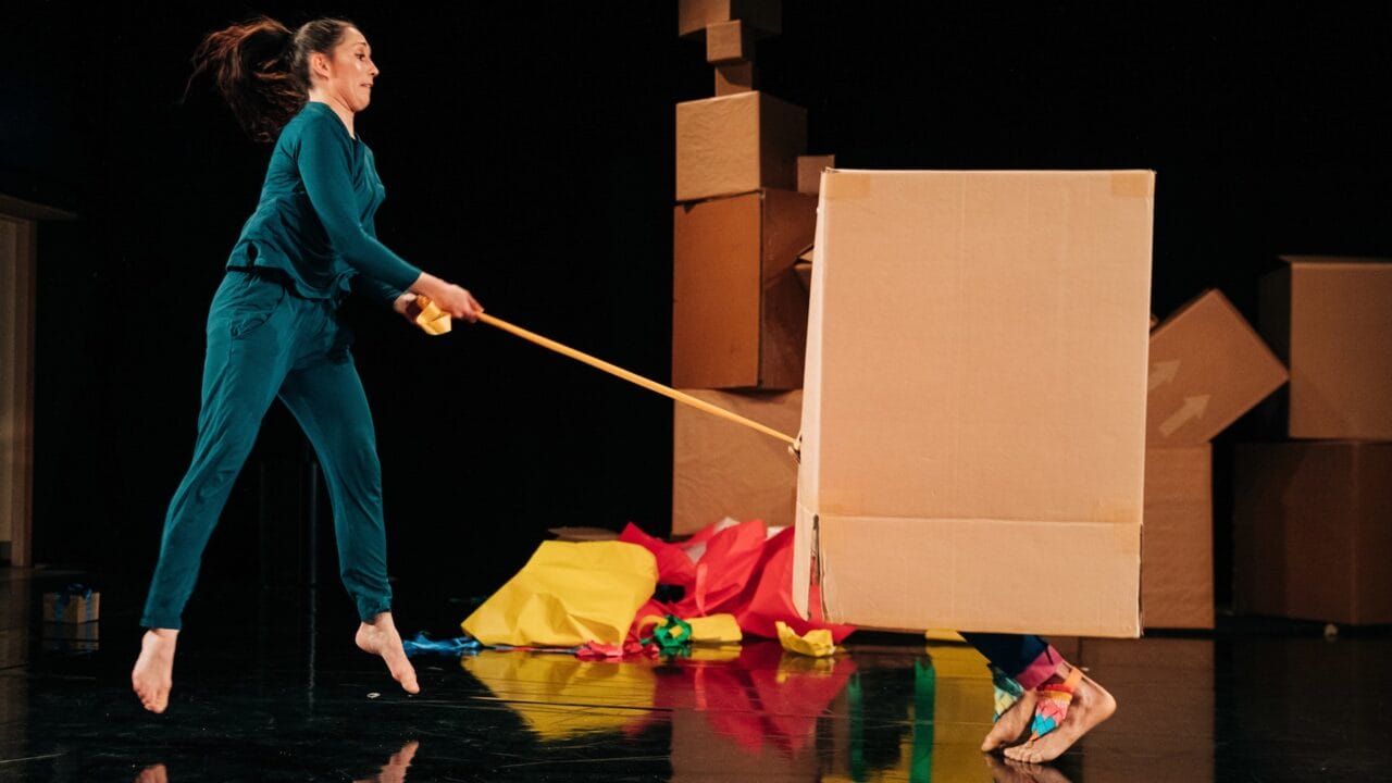 A person in teal jumps while hitting a large cardboard box with a stick; another person is partially hidden inside the box. Crumpled colourful paper is scattered on the floor.