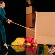 A person in teal jumps while hitting a large cardboard box with a stick; another person is partially hidden inside the box. Crumpled colourful paper is scattered on the floor.