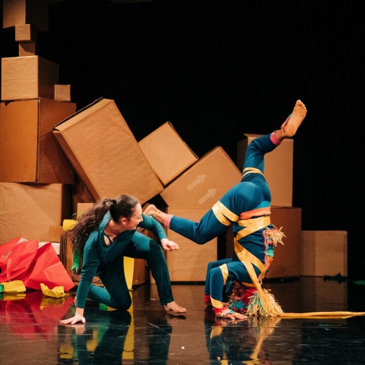 Two performers in colourful costumes pose onstage amid large cardboard boxes, with one person balancing on their head and the other kneeling beside them.