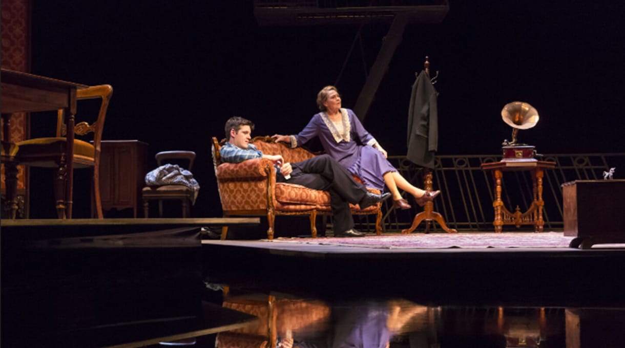A young man and a woman sit on a patterned sofa onstage in a period play set with vintage furniture, a gramophone, and a coat hanging on a stand.