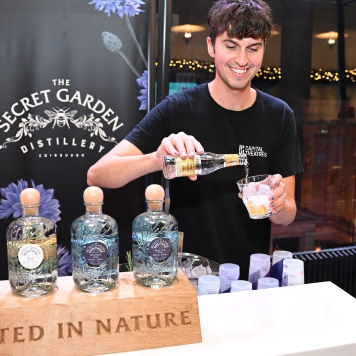 A man pours a drink behind a bar displaying The Secret Garden Distillery gin bottles with a sign reading "Rooted in Nature.