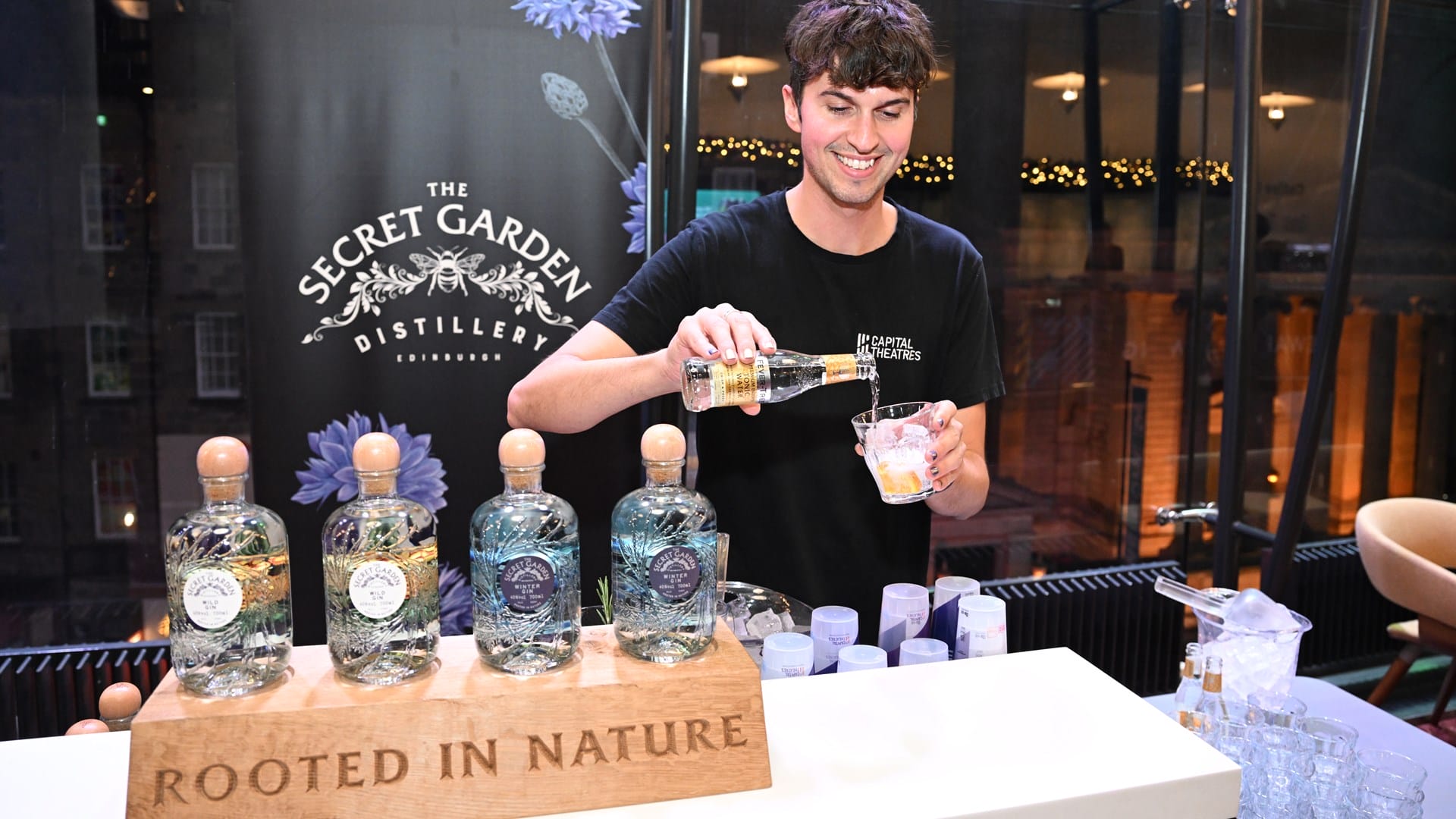 A man pours a drink behind a bar displaying The Secret Garden Distillery gin bottles with a sign reading "Rooted in Nature.