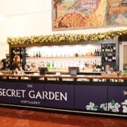A bar counter labelled "The Secret Garden Distillery" with bottles of alcohol on shelves behind, two staff members, and festive decorations above.