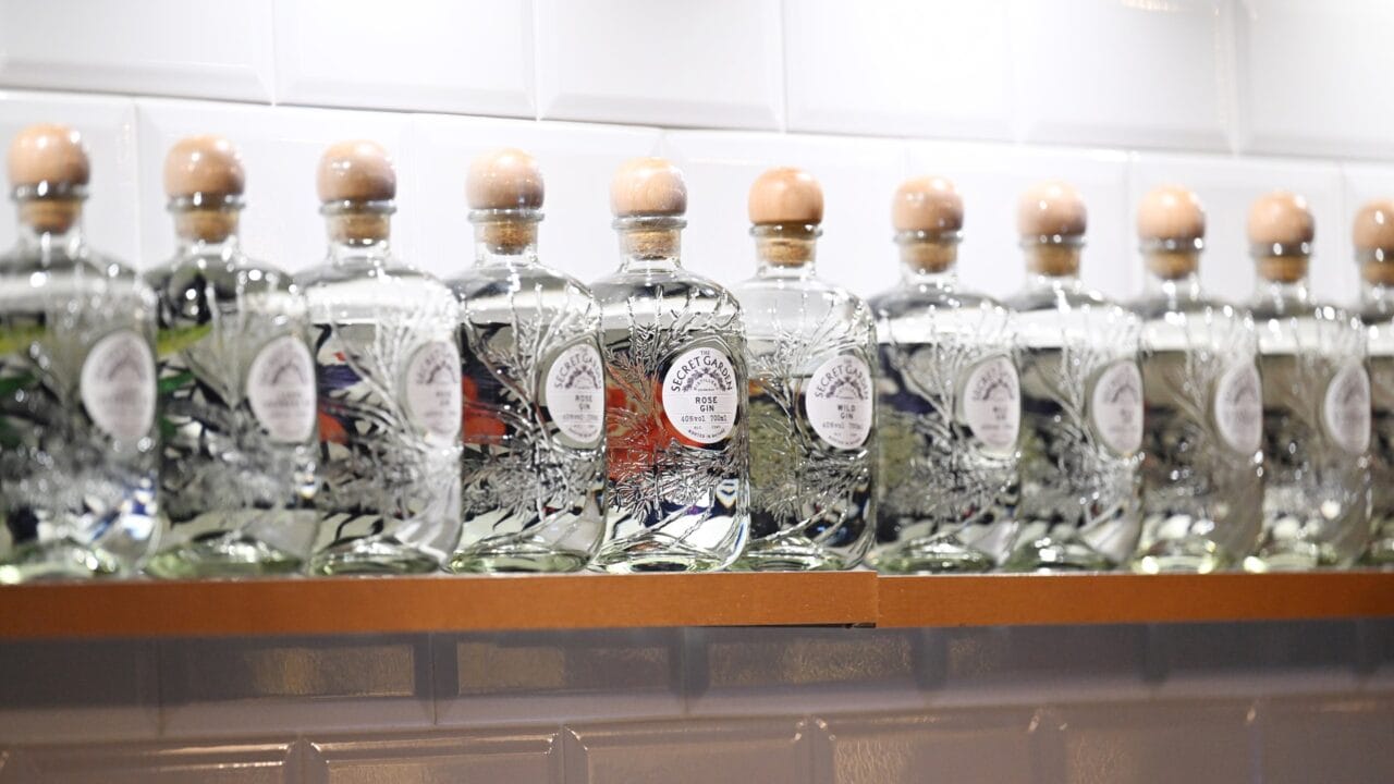 A row of identical glass gin bottles with wooden stoppers displayed on a shelf against a tiled white wall.