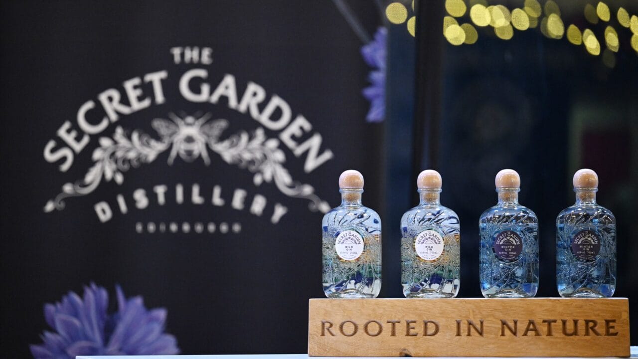Four glass gin bottles from The Secret Garden Distillery are displayed on a wooden stand labelled "Rooted in Nature," with a branded backdrop behind them.