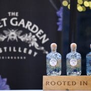 Four glass gin bottles from The Secret Garden Distillery are displayed on a wooden stand labelled "Rooted in Nature," with a branded backdrop behind them.