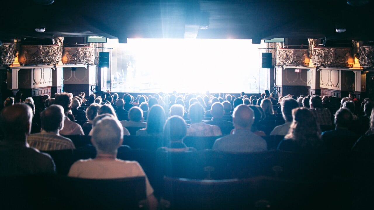 A crowded theater audience sits facing a brightly lit stage or screen, with ornate balcony decorations visible on the sides.