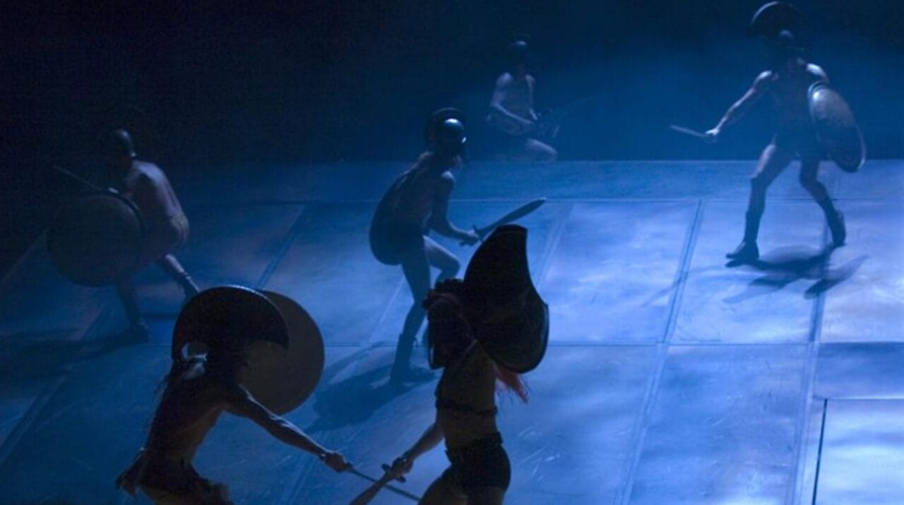 Five people in ancient warrior costumes, holding shields and swords, perform a choreographed battle scene on a dimly lit stage.