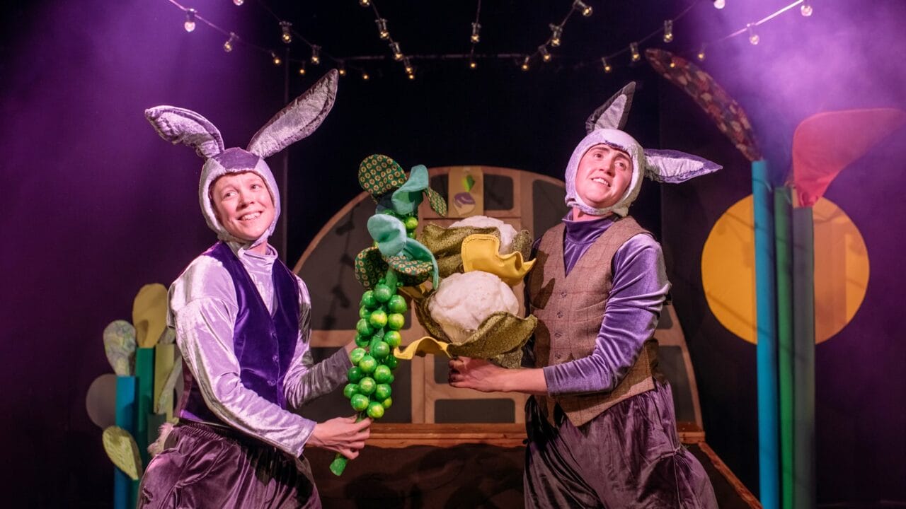 Two people dressed as rabbits hold large plush vegetables on a stage with colorful props and string lights in the background.