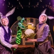 Two people dressed as rabbits hold large plush vegetables on a stage with colorful props and string lights in the background.