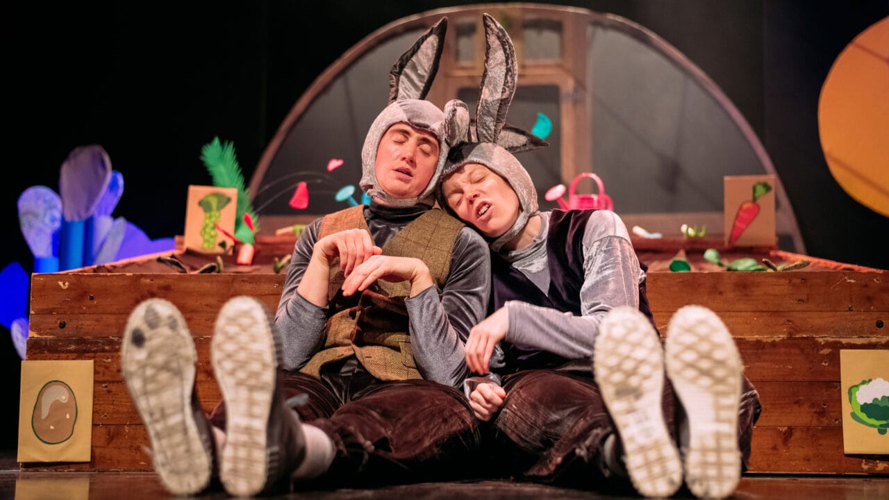 Two people in rabbit costumes sit on the floor, leaning against each other with eyes closed, in front of a colourful stage set featuring vegetables and wooden boxes.