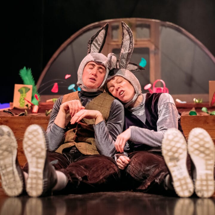 Two people in rabbit costumes sit on the floor, leaning against each other with eyes closed, in front of a colourful stage set featuring vegetables and wooden boxes.