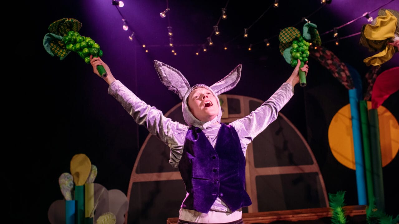 A person in a rabbit costume stands on stage with arms raised, holding green props, under string lights and dramatic purple stage lighting.