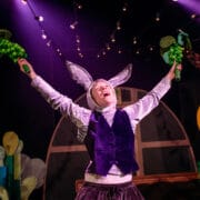 A person in a rabbit costume stands on stage with arms raised, holding green props, under string lights and dramatic purple stage lighting.