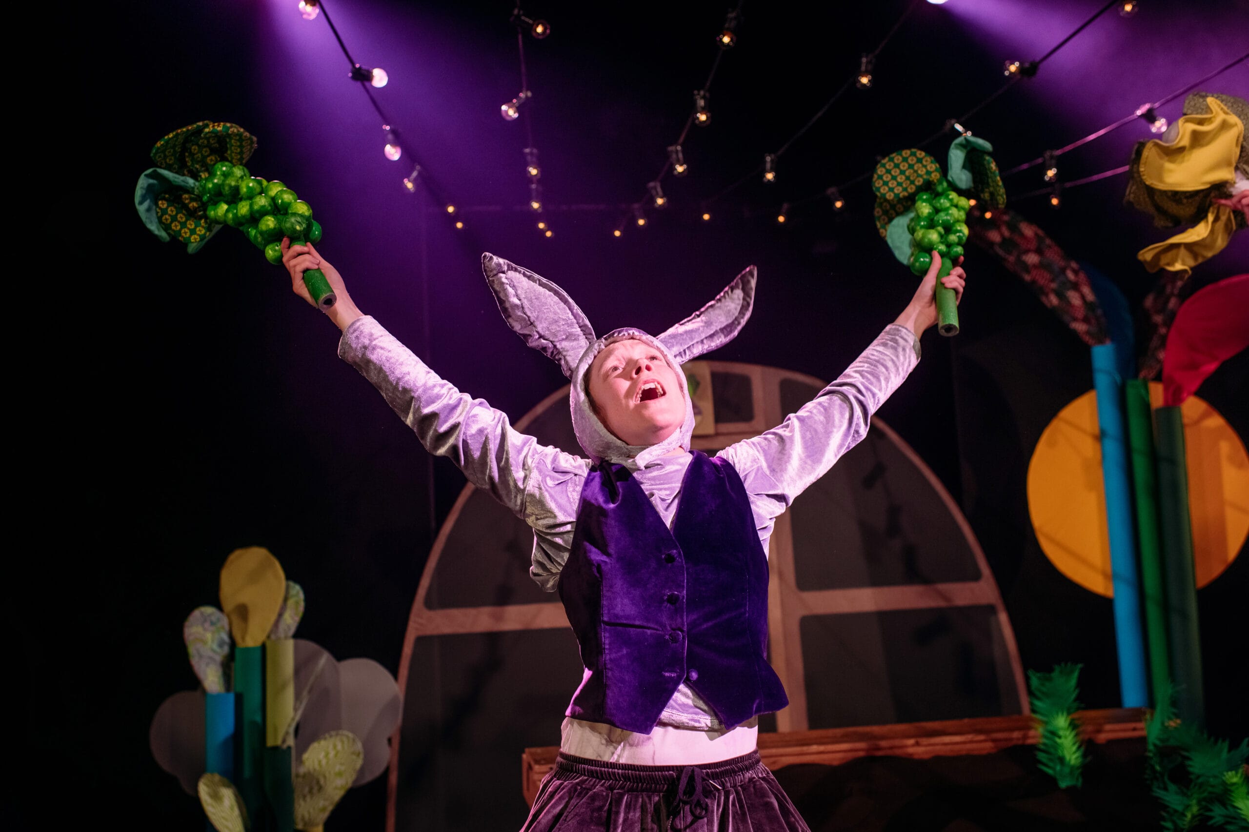 A person in a rabbit costume stands on stage with arms raised, holding green props, under string lights and dramatic purple stage lighting.