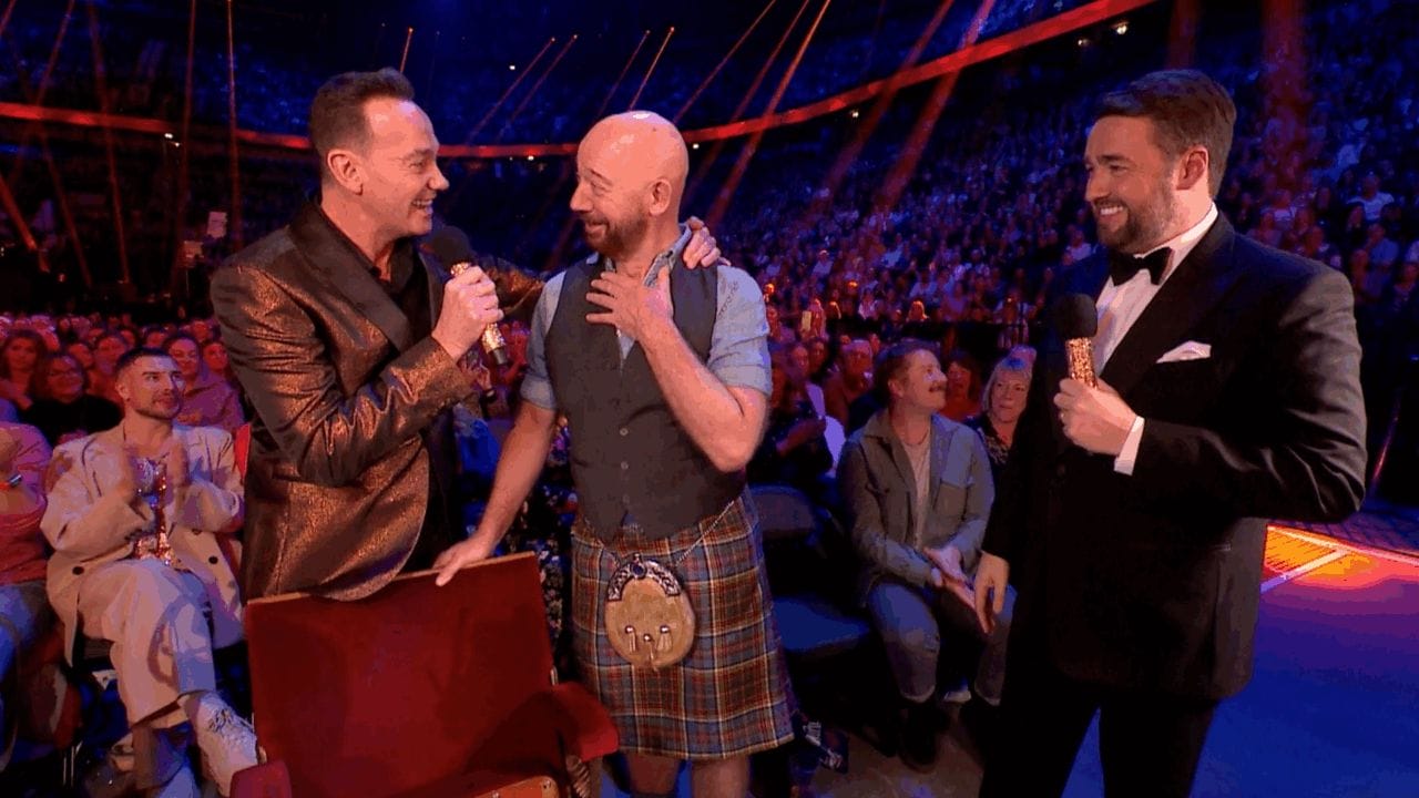 Three men stand on stage, one in a kilt, as they speak into microphones before an applauding audience in a large, brightly lit venue.
