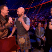 Three men stand on stage, one in a kilt, as they speak into microphones before an applauding audience in a large, brightly lit venue.