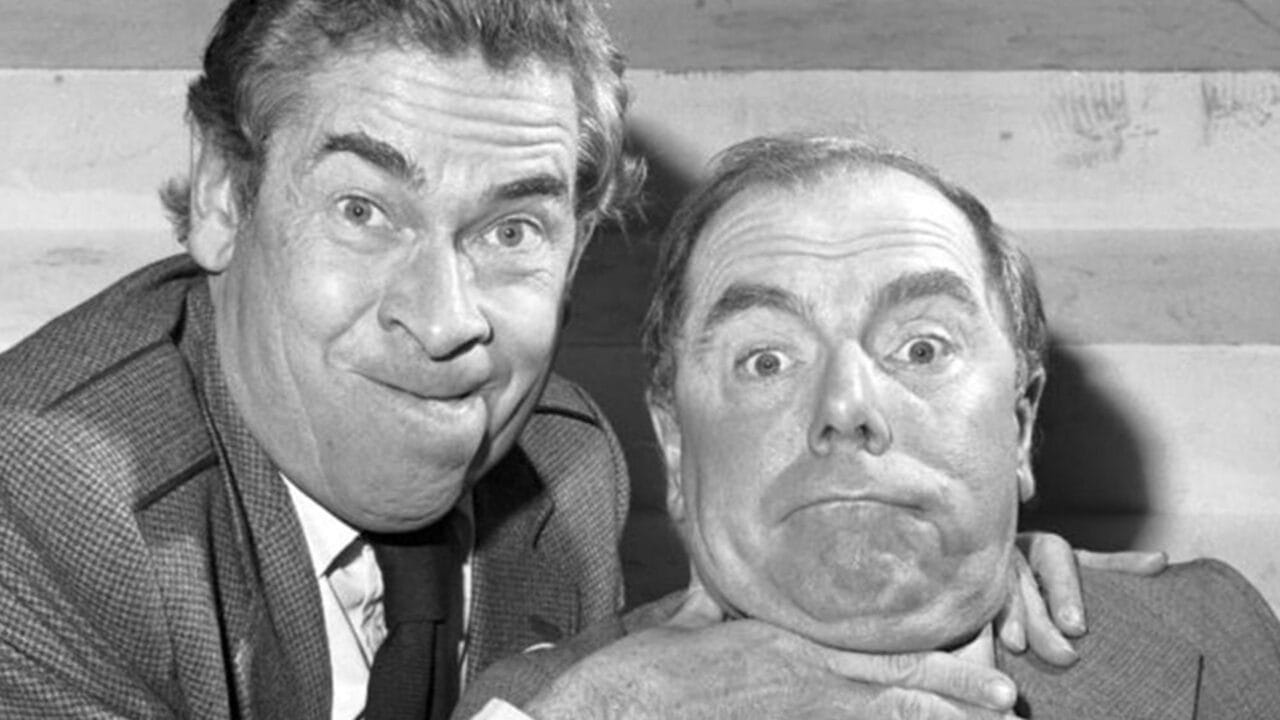 Two men in suits pull exaggerated faces; one man clasps the other’s neck in a playful gesture. The photo is in black and white.