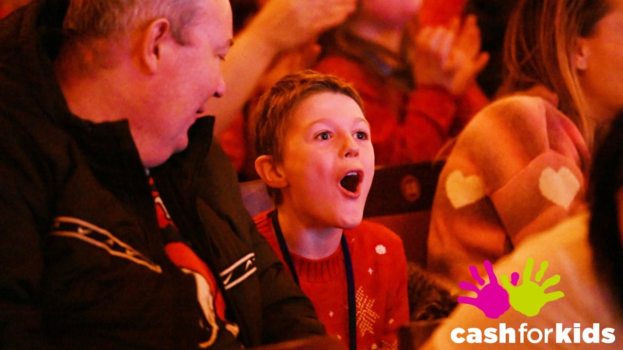 A child with an excited expression sits among adults at an event. "Cash for Kids" logo with colorful handprints is in the lower right corner.