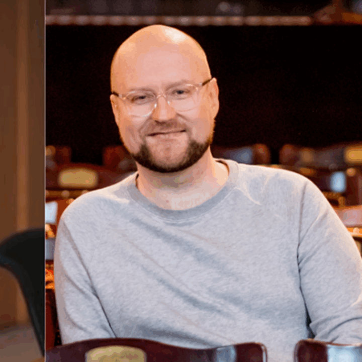 A woman with long brown hair in a blue dress sits smiling next to a man with glasses and a beard wearing a grey sweater, both indoors.