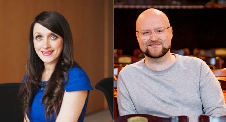 A woman with long brown hair in a blue dress sits smiling next to a man with glasses and a beard wearing a grey sweater, both indoors.