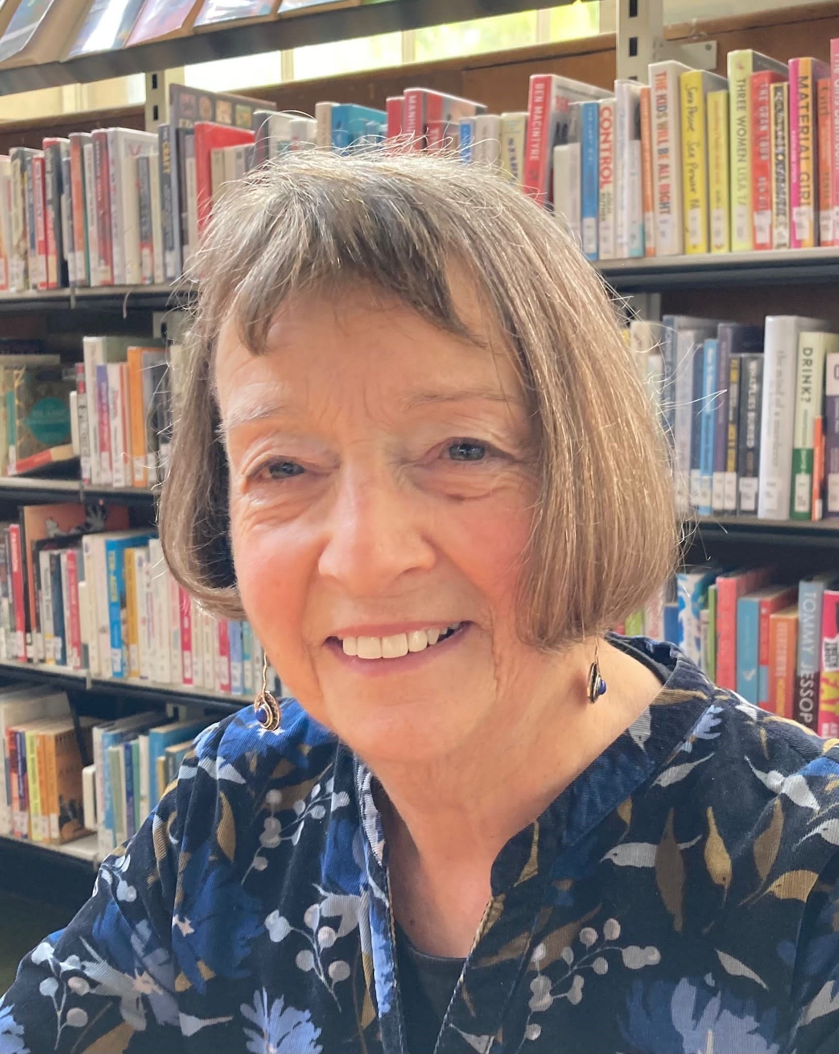 An older woman with short brown hair smiles at the camera while sitting in front of bookshelves in a library.