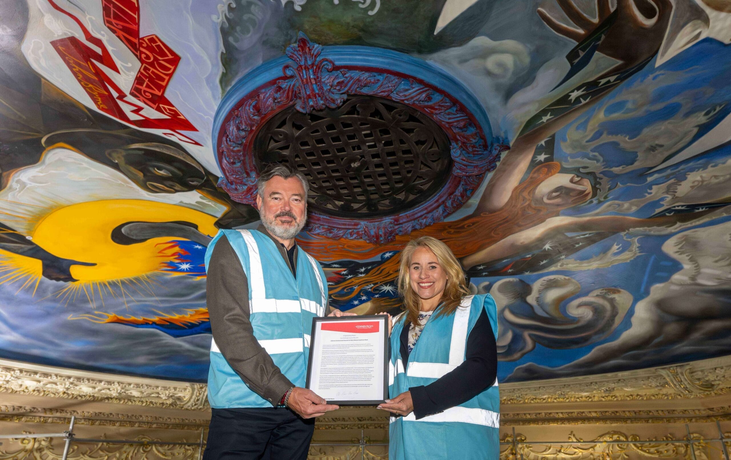 Grant Stott and Councillor Munro hold a framed document while standing under the King's Theatre ceiling mural.