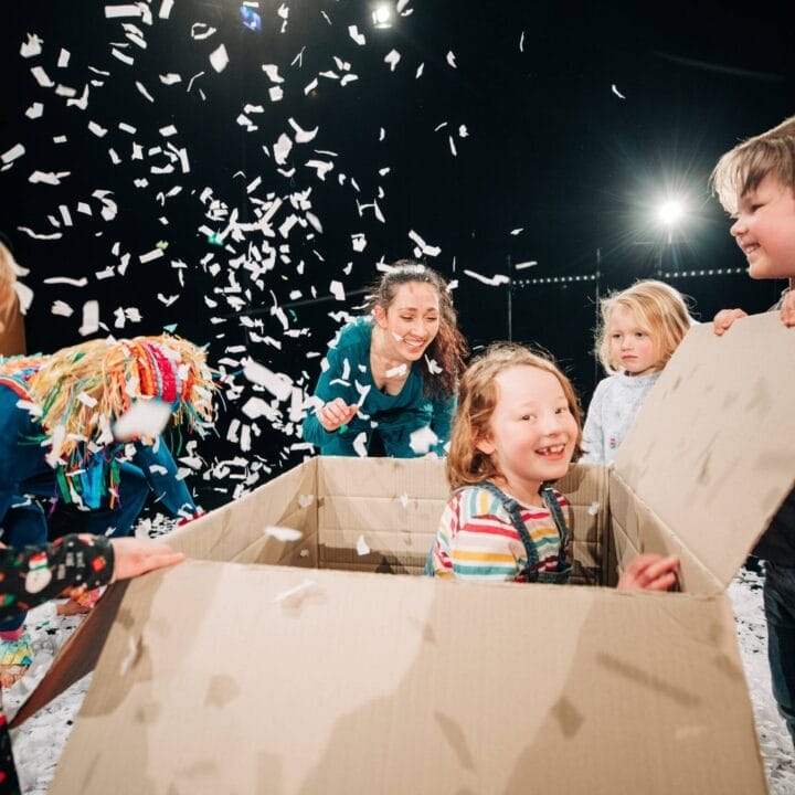 Children play with confetti and a large cardboard box indoors, with one child sitting inside the box and other Wee Ones gathered around, smiling and laughing.