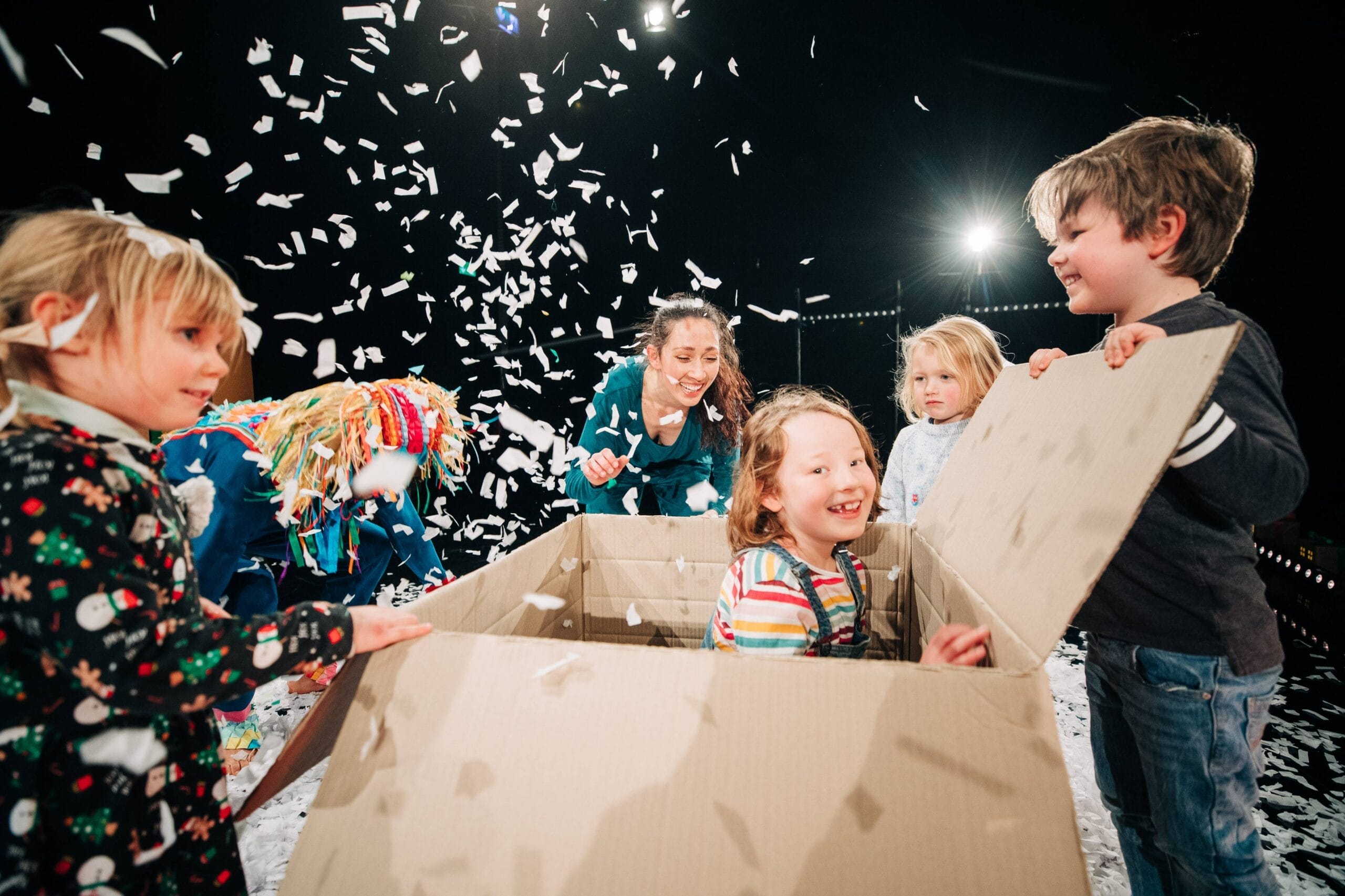 Children play with confetti and a large cardboard box indoors, with one child sitting inside the box and other Wee Ones gathered around, smiling and laughing.