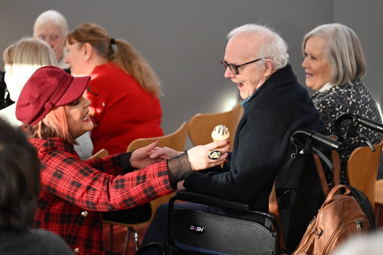 A woman in a red plaid jacket hands a treat to an elderly man in a wheelchair.
