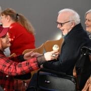 A woman in a red plaid jacket hands a treat to an elderly man in a wheelchair.