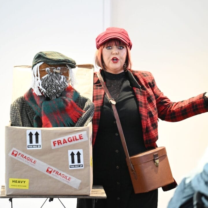 A woman in a red plaid jacket and hat stands beside a large box labelled "FRAGILE,". Inside, a figure with a knitted face, hat, and scarf awaits.