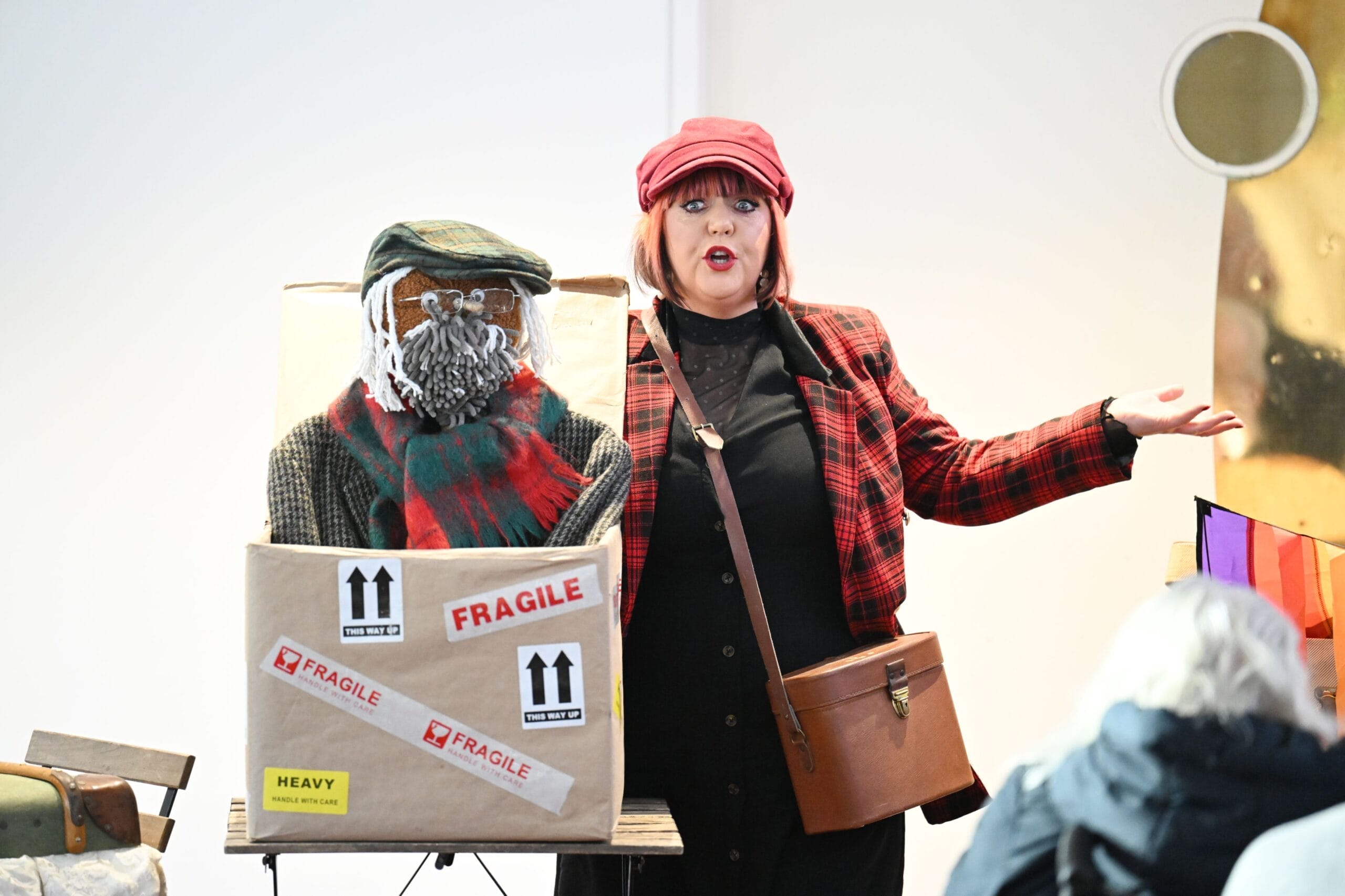 A woman in a red plaid jacket and hat stands beside a large box labelled "FRAGILE,". Inside, a figure with a knitted face, hat, and scarf awaits.