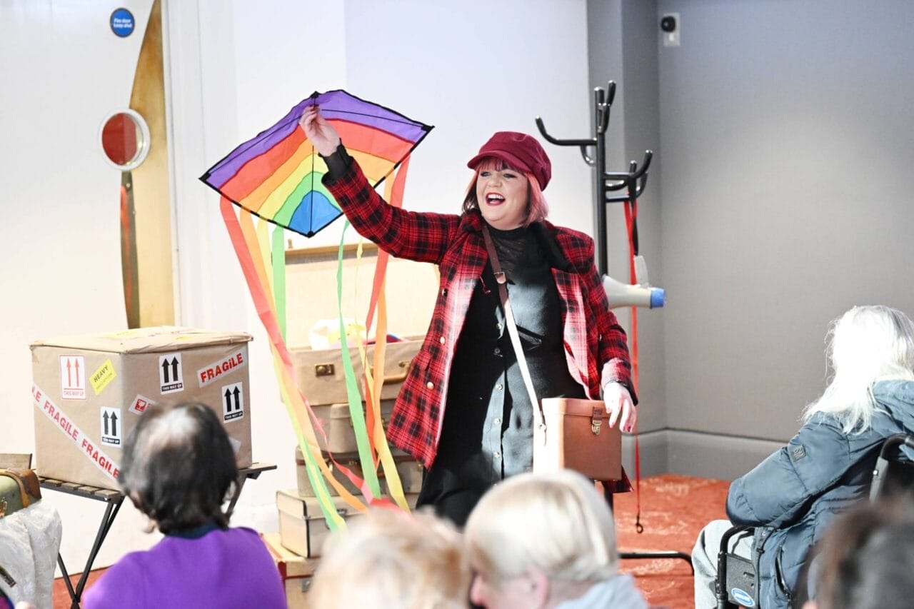 A woman in a red plaid jacket and hat is flying a rainbow kite while an audience watches on.