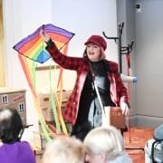 A woman in a red plaid jacket and hat is flying a rainbow kite while an audience watches on.