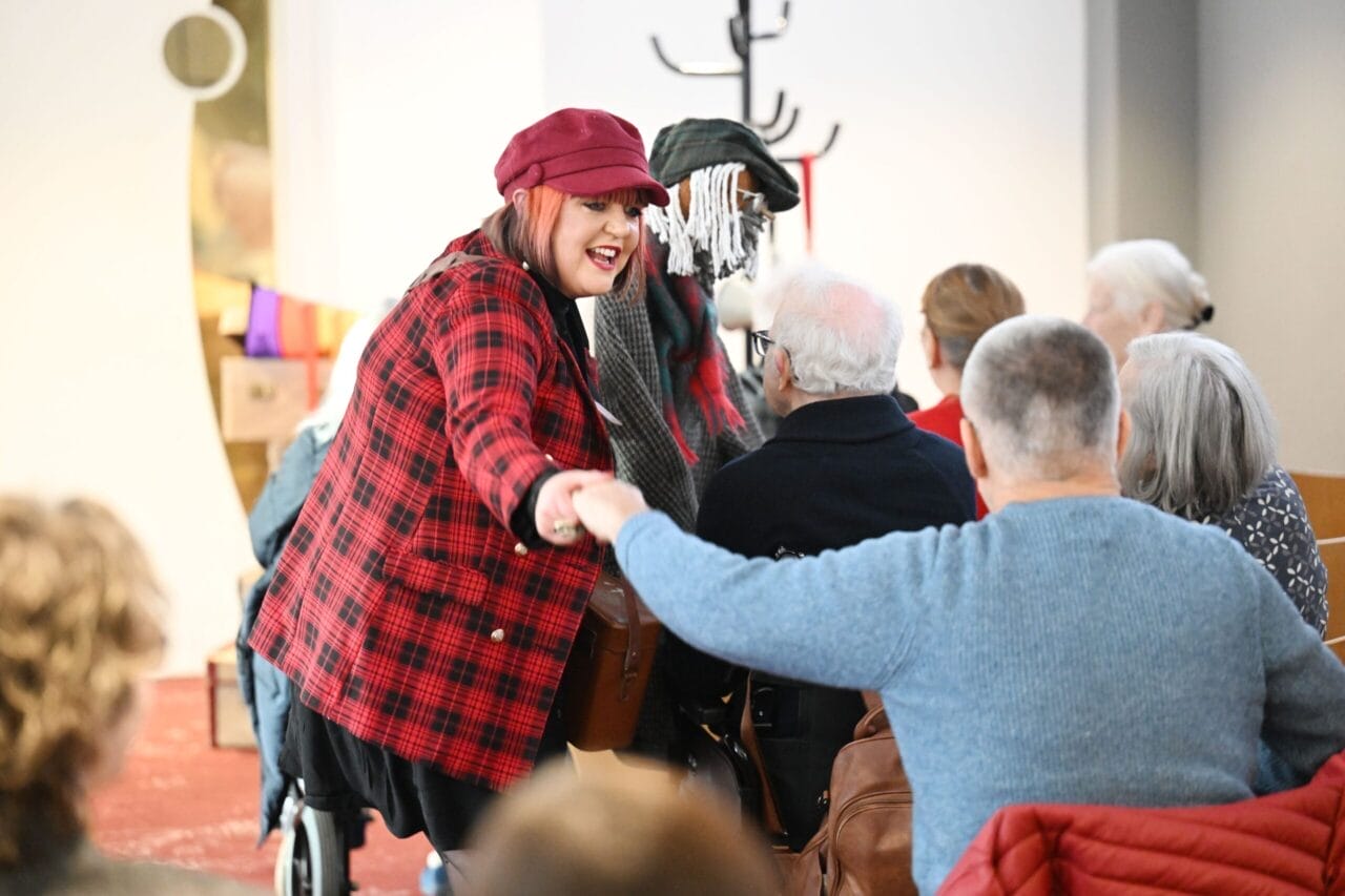 A woman in a red plaid jacket and hat holds the hand of an audience member.