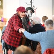A woman in a red plaid jacket and hat holds the hand of an audience member.