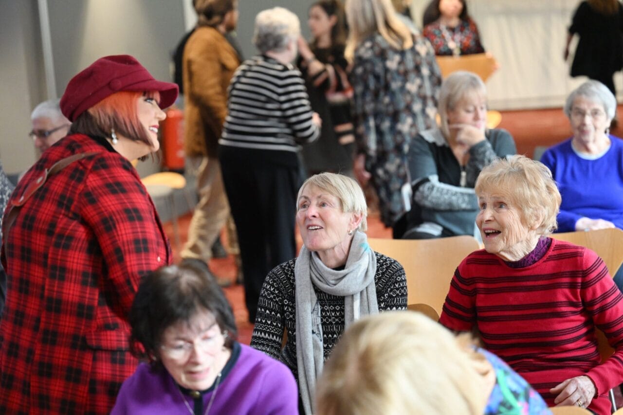 A group of older adults sit and chat in a bright room while others stand and converse in the background.