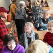 A group of older adults sit and chat in a bright room while others stand and converse in the background.