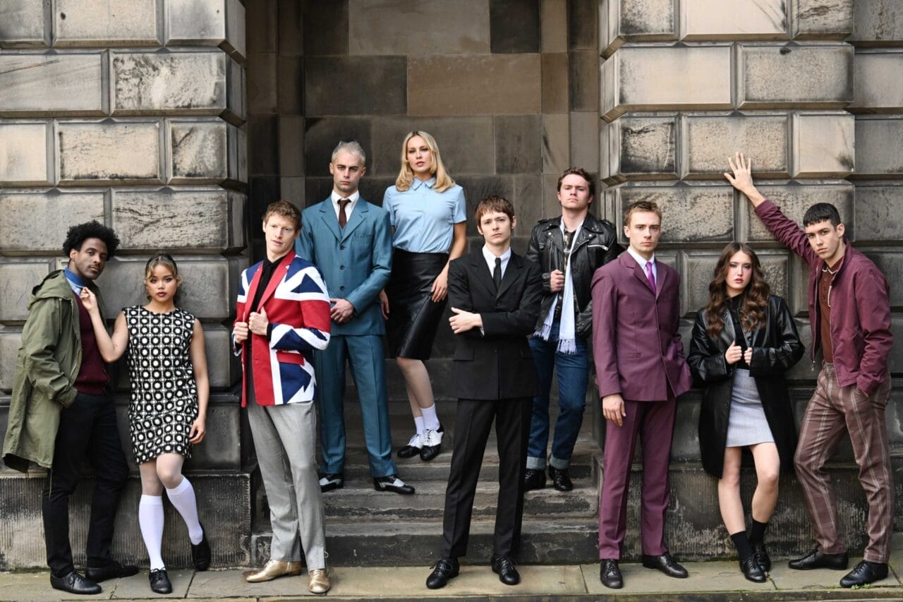 Ten young adults in 1960s-inspired outfits pose in front of a stone building.