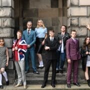 Ten young adults in 1960s-inspired outfits pose in front of a stone building.