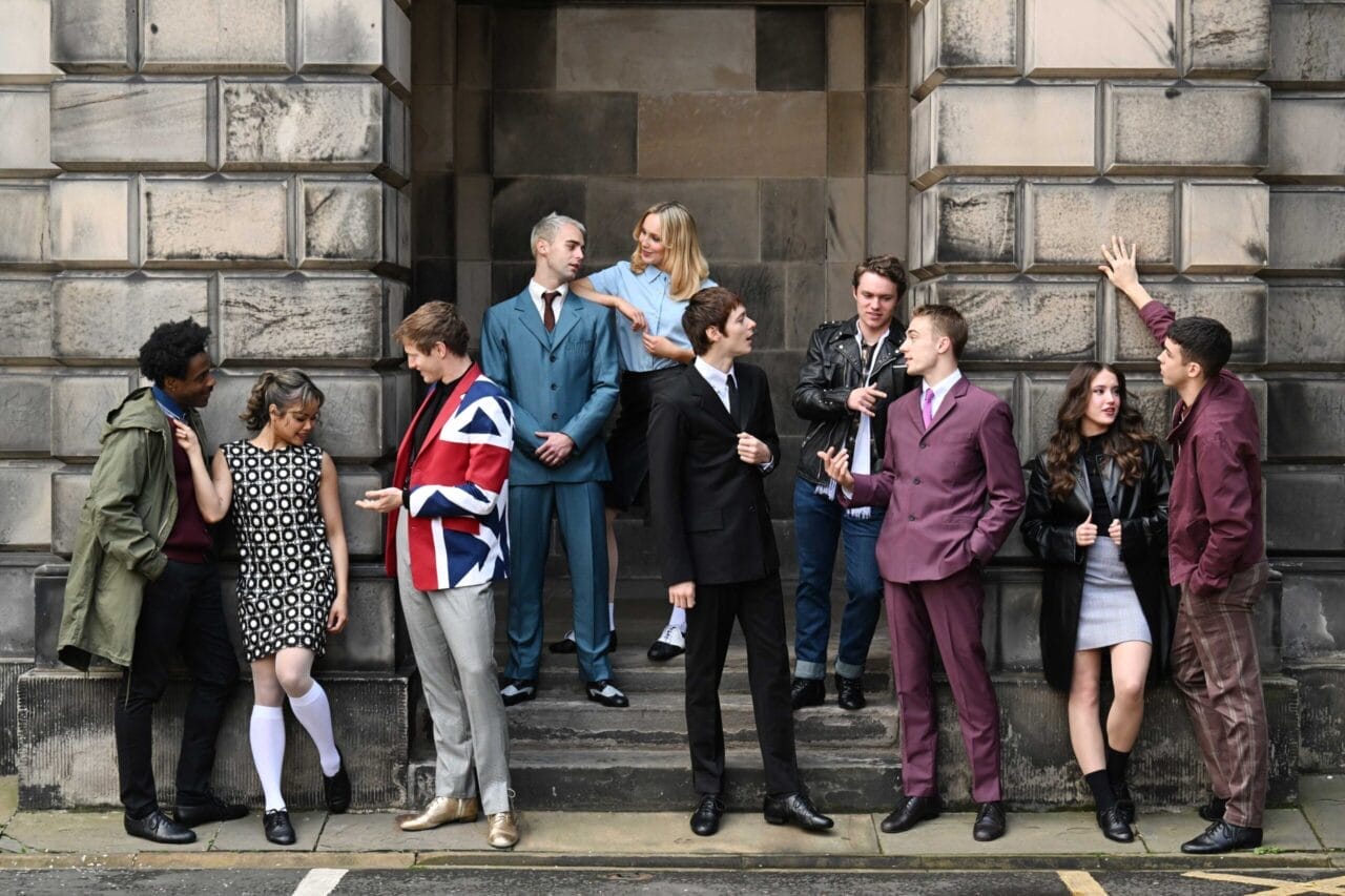 A group of young adults in various retro-style outfits stand and converse outside a stone building, some on steps and others on the pavement.
