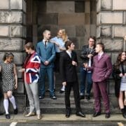 A group of young adults in various retro-style outfits stand and converse outside a stone building, some on steps and others on the pavement.