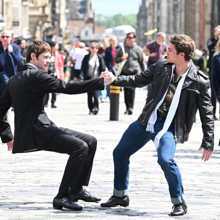 Two young men, one in a suit and one in a leather jacket, hold hands and perform a playful Mod Ballet dance move on a busy cobblestone street filled with pedestrians.