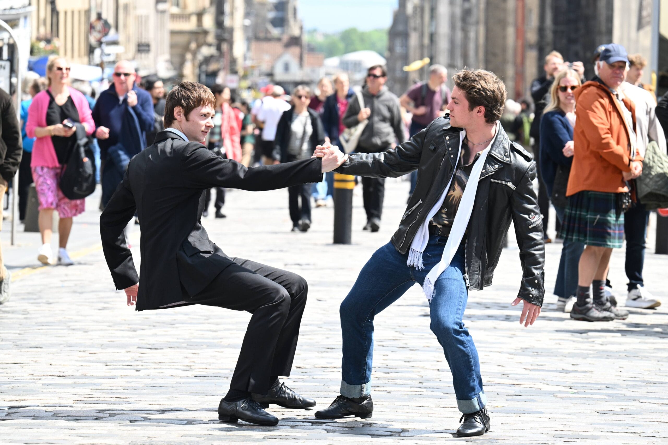 Two young men, one in a suit and one in a leather jacket, hold hands and perform a playful Mod Ballet dance move on a busy cobblestone street filled with pedestrians.