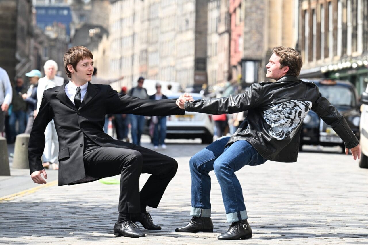 Two young men balance by holding hands while leaning back on a cobblestone street.