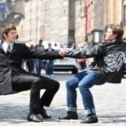 Two young men balance by holding hands while leaning back on a cobblestone street.