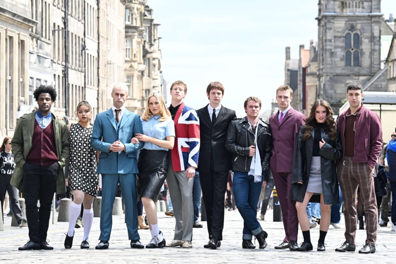 A group of nine young adults stand in a row on a city street, dressed in a mix of retro and modern clothing, channeling Mod Ballet flair, with historic buildings visible in the background.